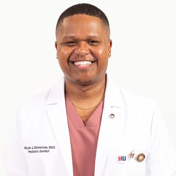Smiling pediatric dentist in a white coat with the name Micah J Dickerson DDS and an HU badge on a white background Smiling pediatric dentist in a white coat with the name Micah J. Dickerson, DDS, and an HU badge on a white background.